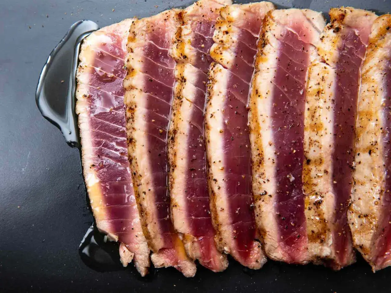 Skivor av perfekt grillad tonfisk, kryddad med peppar och serverad med olja på en mörk yta.