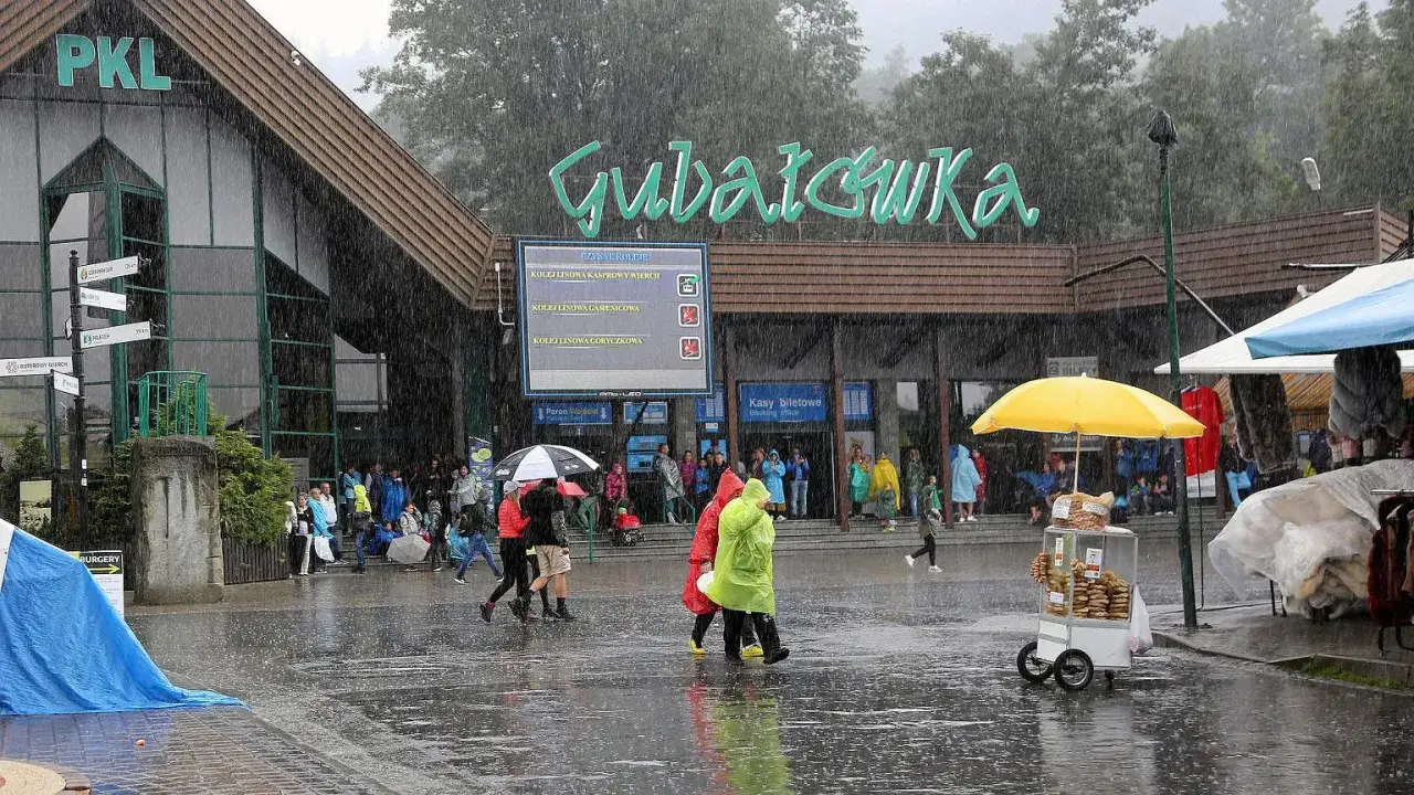 Ludzie w kolorowych płaszczach przeciwdeszczowych i pod parasolami spieszą się pod Gubał&oacute;wkę w deszczu. Mokry chodnik odbija światła.