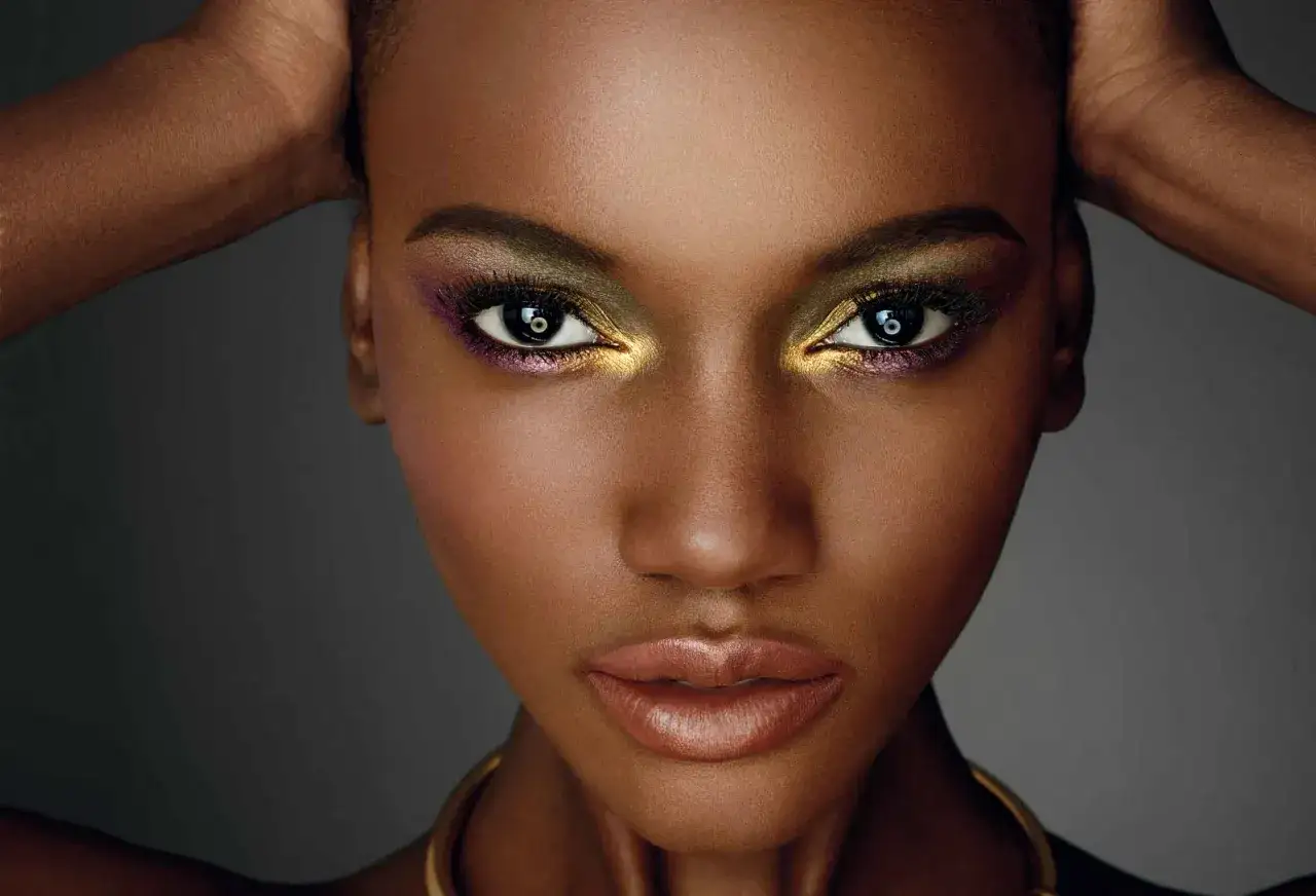 Un maquillage soirée doré et audacieux illumine le regard d'une femme, rehaussé de touches violettes. Ses lèvres sont subtilement glossées.