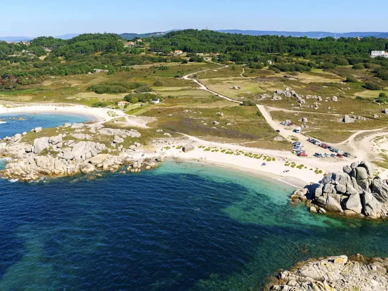 Playa con rocas y un grove de pinos al fondo. El mar turquesa ba&ntilde;a la arena blanca, con coches aparcados cerca.