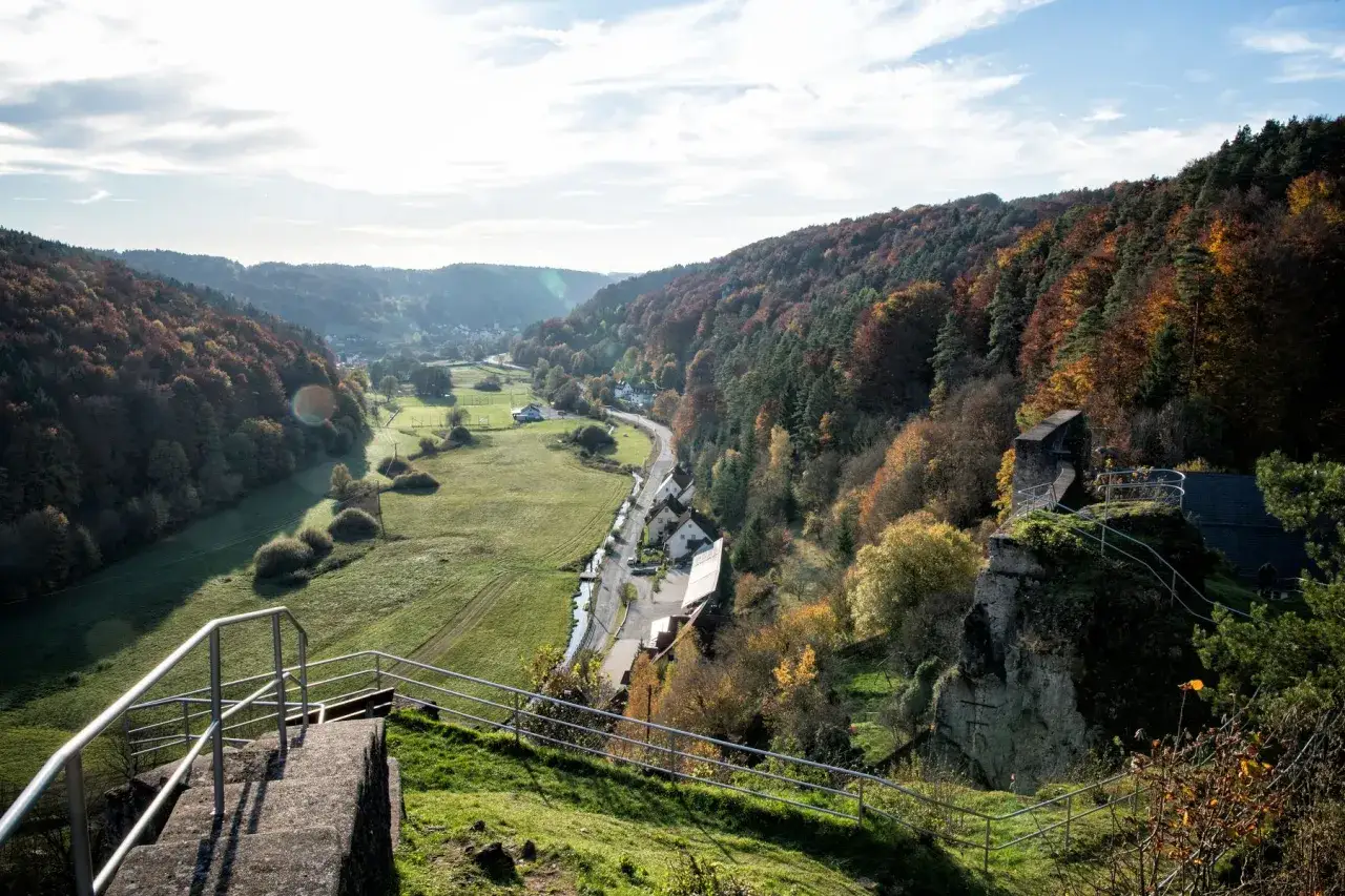 Blick auf die Burgruine Wolfsberg im Herbst. Ein malerisches Tal mit bunten Wäldern, grünen Wiesen und einem kleinen Dorf.