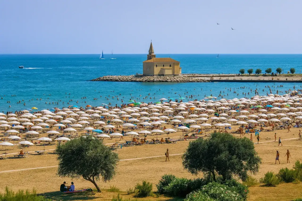 Vue panoramique de la **Caorle plage** bond&eacute;e, avec des rang&eacute;es de parasols blancs, des baigneurs dans la mer bleue et une petite &eacute;glise sur une jet&eacute;e.