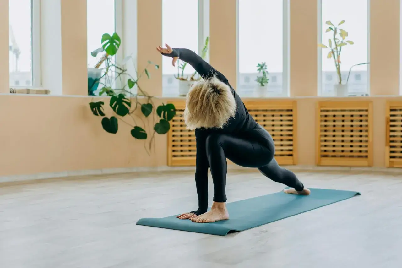 Kvinna i svart tr&auml;ningskl&auml;dsel utf&ouml;r en yoga&ouml;vning p&aring; en bl&aring; matta. Hon str&auml;cker ut armen &ouml;ver huvudet, en pose som fr&auml;mjar yoga avslappning.