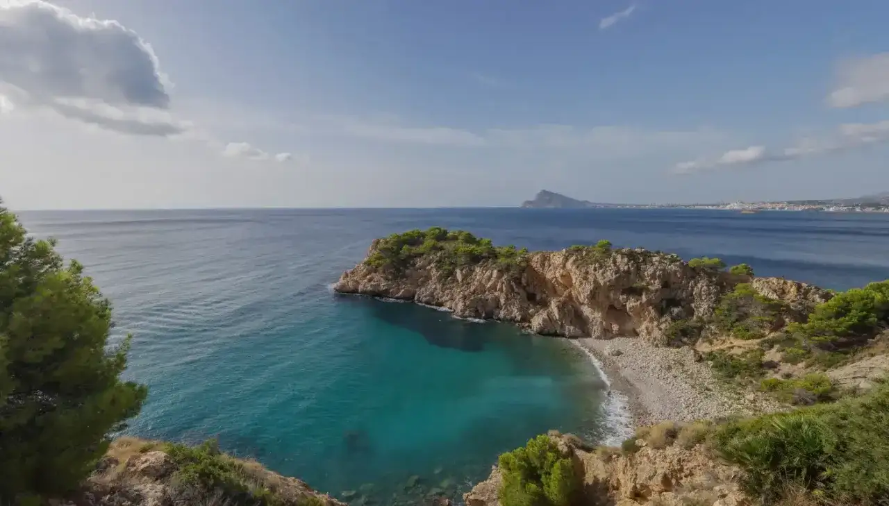 Vista de una cala con aguas turquesas y rocas cubiertas de vegetación. Al fondo, el mar azul se extiende hasta la costa con edificios.