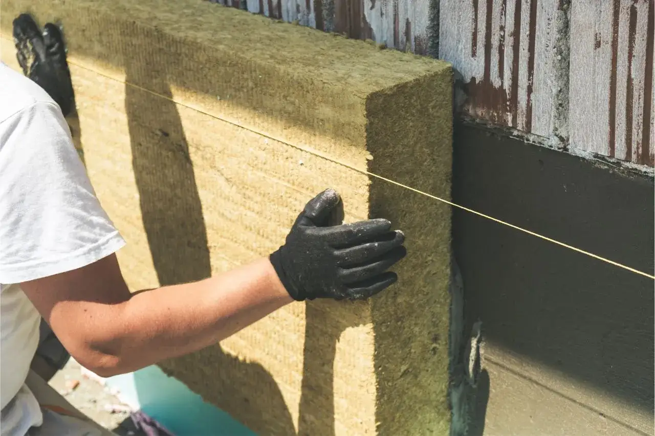 Colocación de aislamiento en vivienda unifamiliar. Un trabajador con guantes negros ajusta un panel de lana de roca contra la pared exterior.
