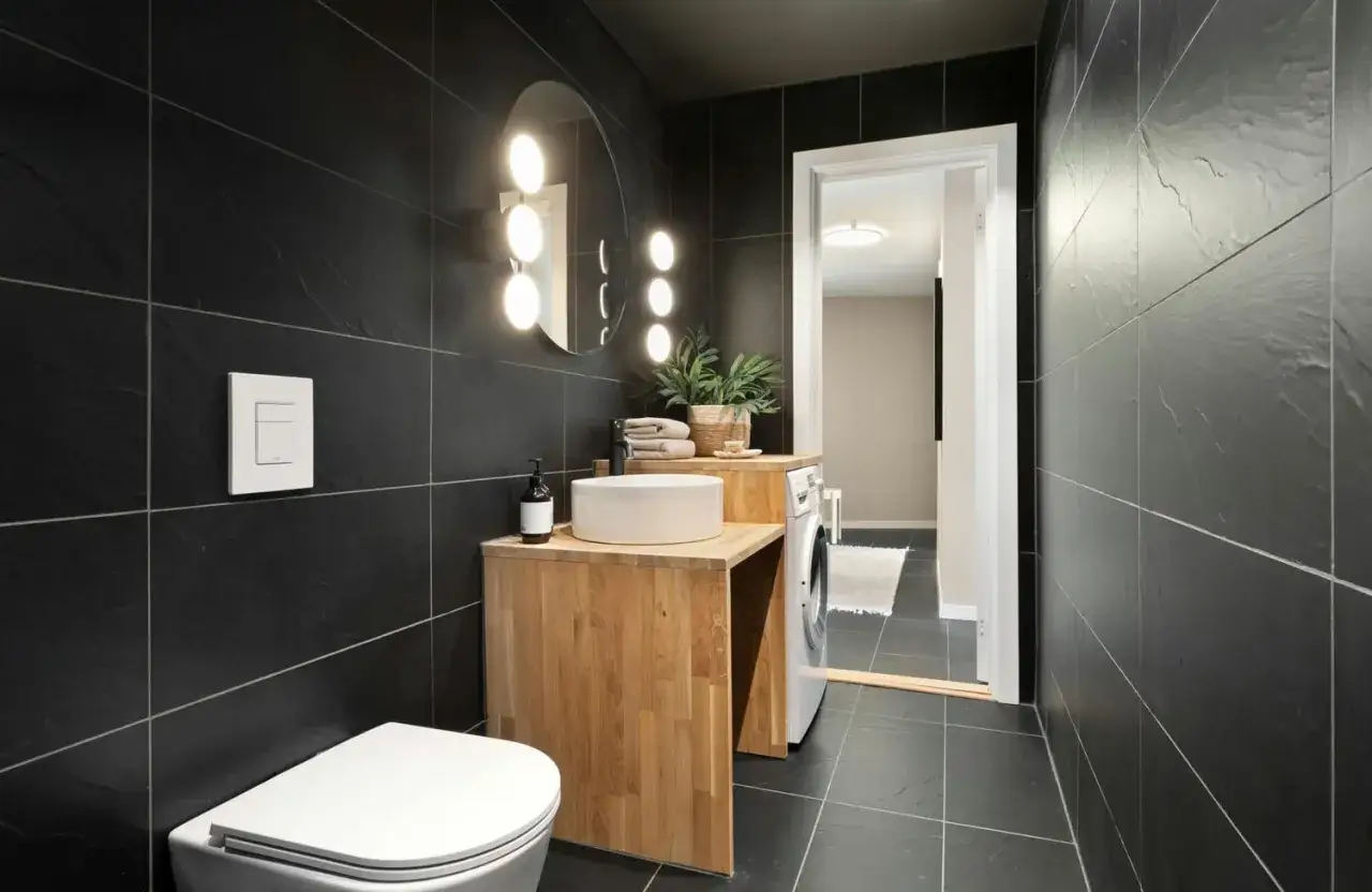 Modern bathroom with black tile and brown grout, featuring a white toilet, wooden vanity with a sink, and a washing machine.