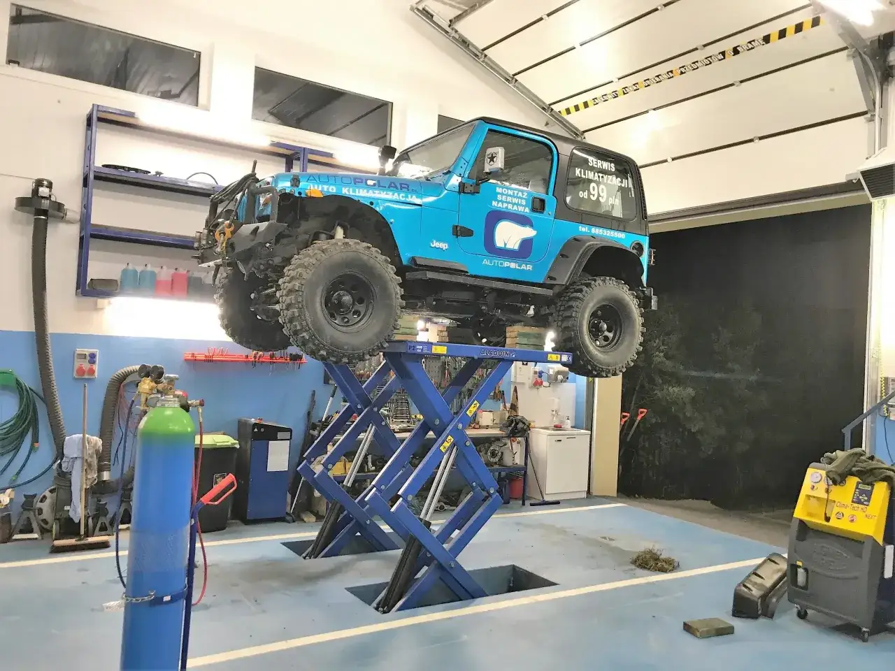Niebieski Jeep na podnośniku, obok żółte urządzenie do płukania klimatyzacji.