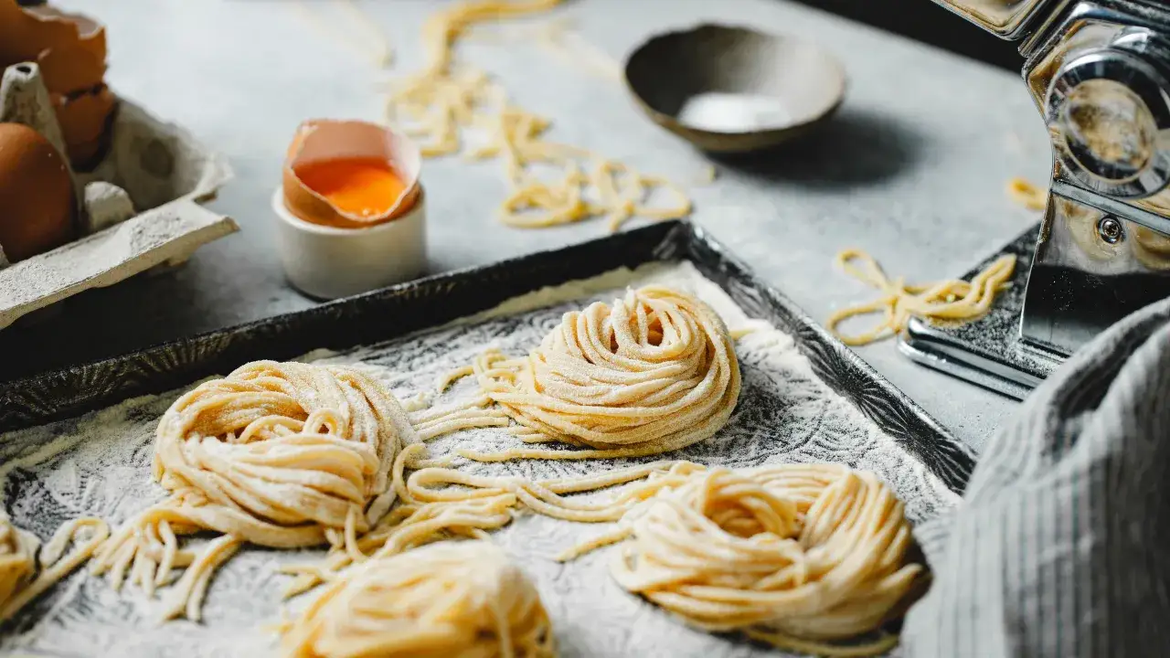 Nidi di pasta fatta in casa, freschi e pronti per essere cucinati, con uova e una macchina per la pasta sullo sfondo.