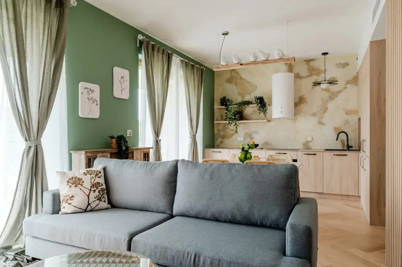 Przytulne, **małe mieszkanie w stylu boho**. Szara kanapa, zielona ściana, drewniane meble i rośliny tworzą relaksującą atmosferę.