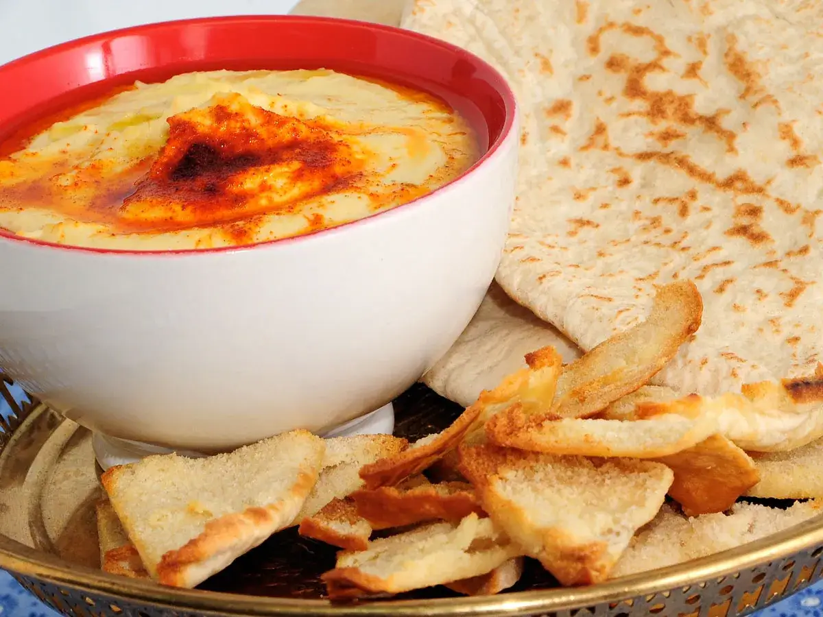 Hummus receta tradicional servido en un cuenco blanco con borde rojo, adornado con pimentón y aceite de oliva. Acompañado de pan de pita y tostadas crujientes.