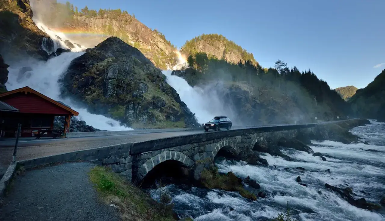 Un coche cruza un puente de piedra sobre un r&iacute;o caudaloso, con una cascada y un arco&iacute;ris al fondo. Una aventura de noruega en coche.