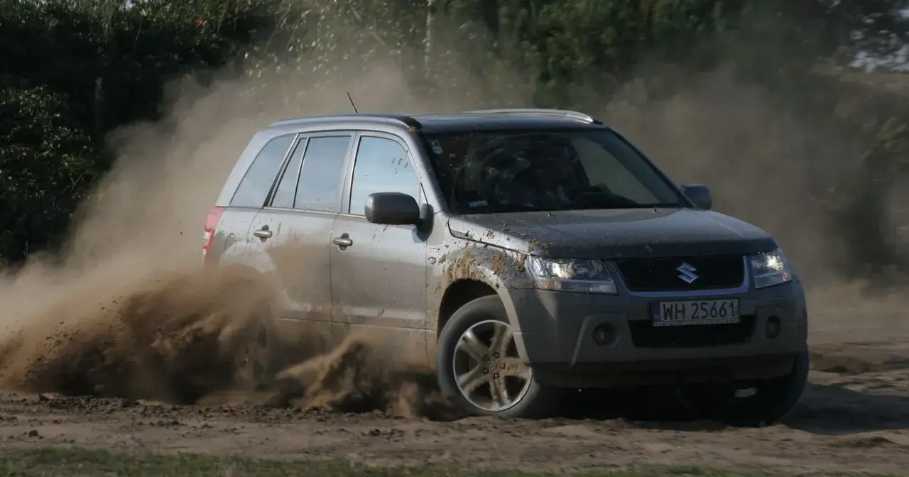 Suzuki Grand Vitara wjeżdża w piasek, wzbijając tumany kurzu.
