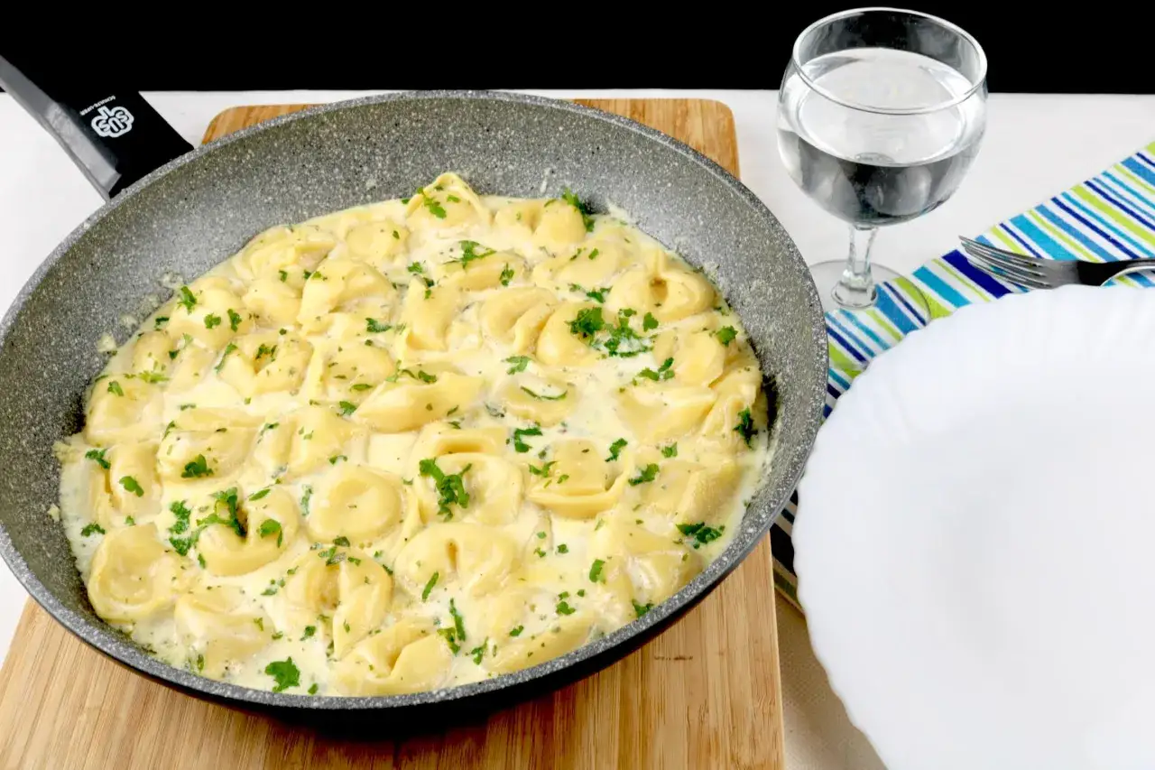 Cremige Tortellini in einer Pfanne, garniert mit Petersilie. Perfekt für schnelle Tortellini Rezepte. Daneben ein Glas Wasser und ein Teller.