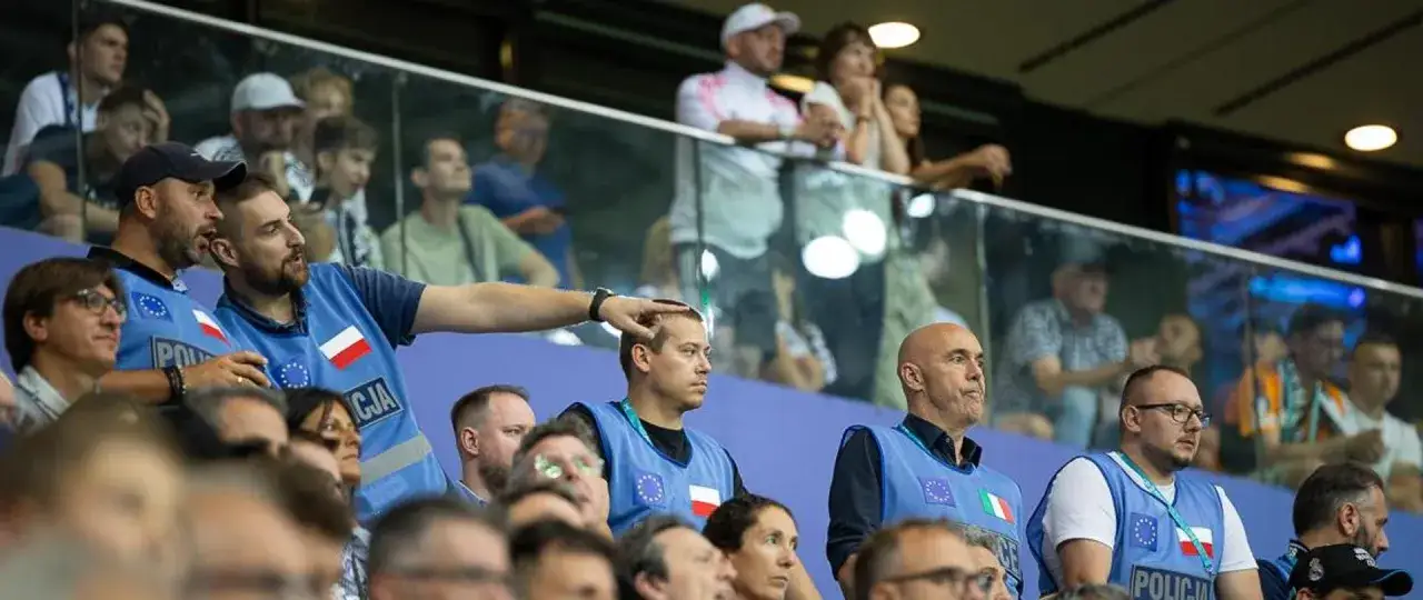 Ochrona w kamizelkach z napisem POLICJA pilnuje porządku wśród kibiców. Pseudokibice w tłumie, niektórzy wpatrzeni w trybuny.
