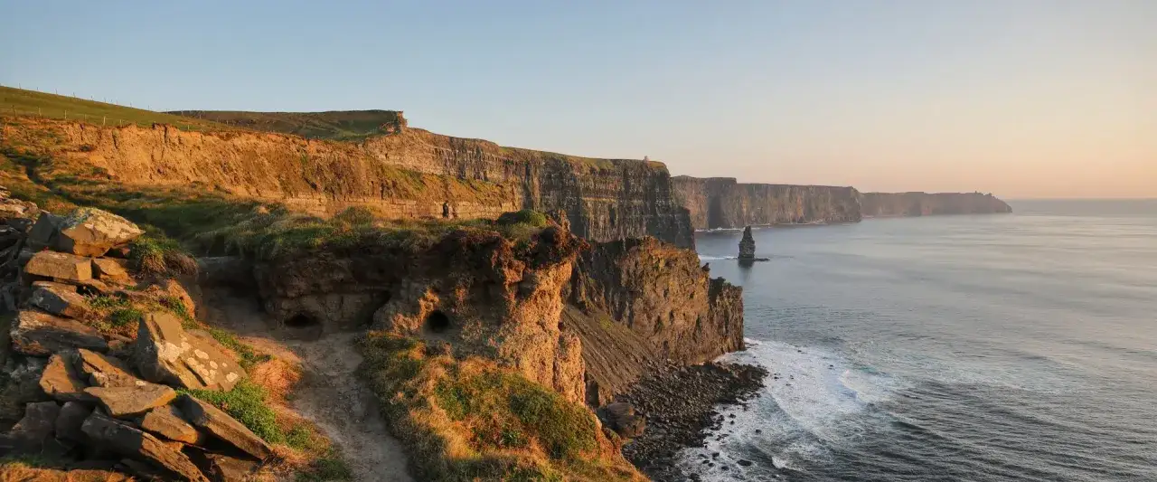 Falaises spectaculaires d'Irlande au coucher du soleil, avec des vagues s'écrasant sur les rochers et un ciel aux teintes chaudes.