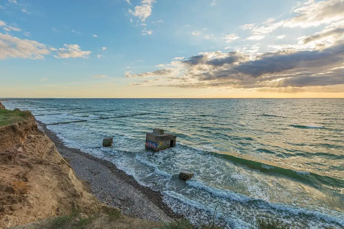 Ein alter Bunker am Strand, umspült von Wellen. Die Sonne bricht durch Wolken und beleuchtet das Meer.