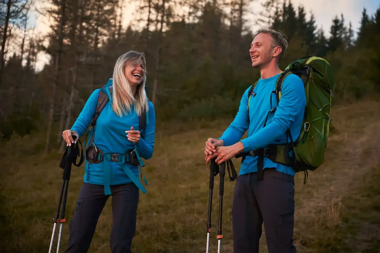 Para wybrać, jak się ubrać w góry, spójrz na parę turystów w niebieskich bluzach i ciemnych spodniach, z plecakami i kijkami trekkingowymi, śmiejących się na tle lasu.