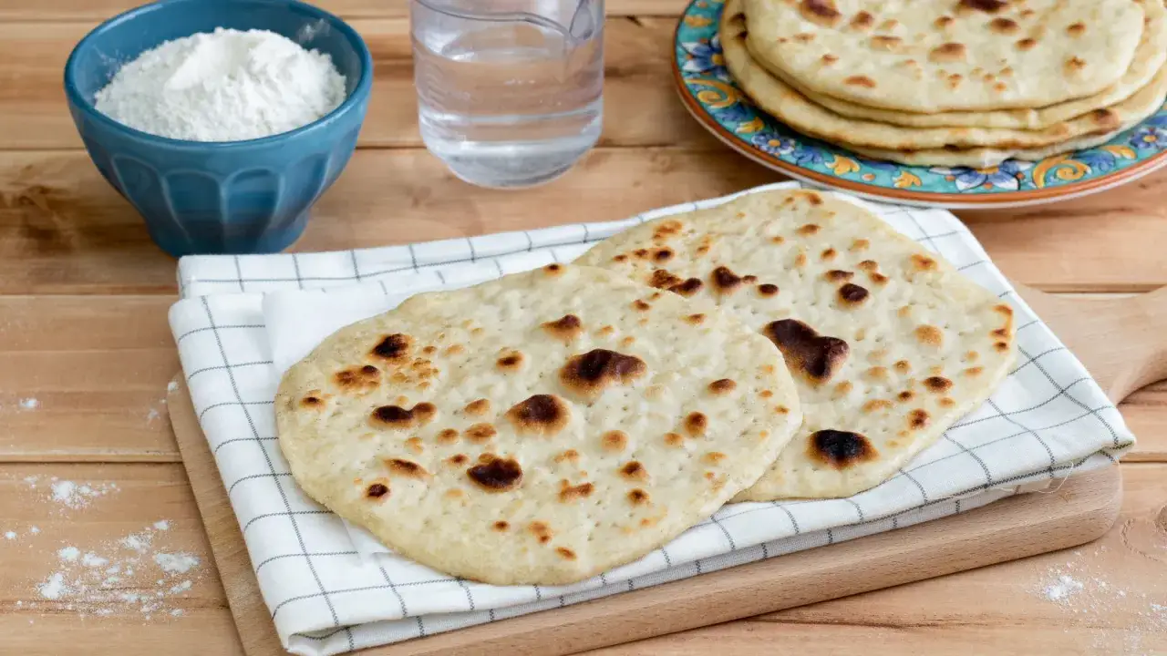 Due focacce dorate, cotte alla perfezione, sono il cuore di questa immagine. Accanto, una ciotola di farina e un bicchiere d'acqua suggeriscono la preparazione casalinga del pane senza lievito.