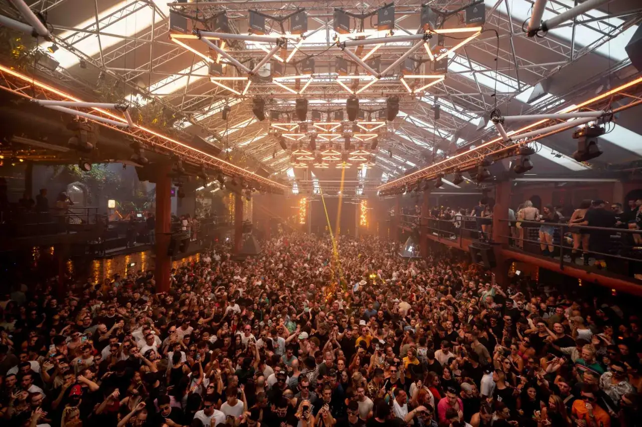 Folla immensa in un locale con luci arancioni e struttura metallica, tipico degli eventi Amnesia Ibiza.