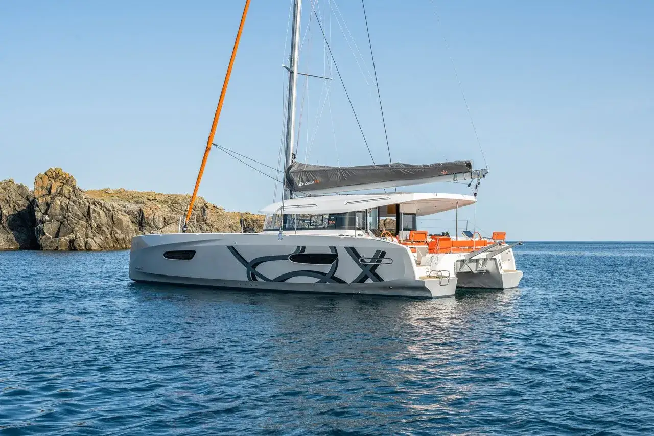 Catamarano Excess 11 con vele grigie e dettagli arancioni, attraccato vicino a scogli rocciosi sotto un cielo azzurro.