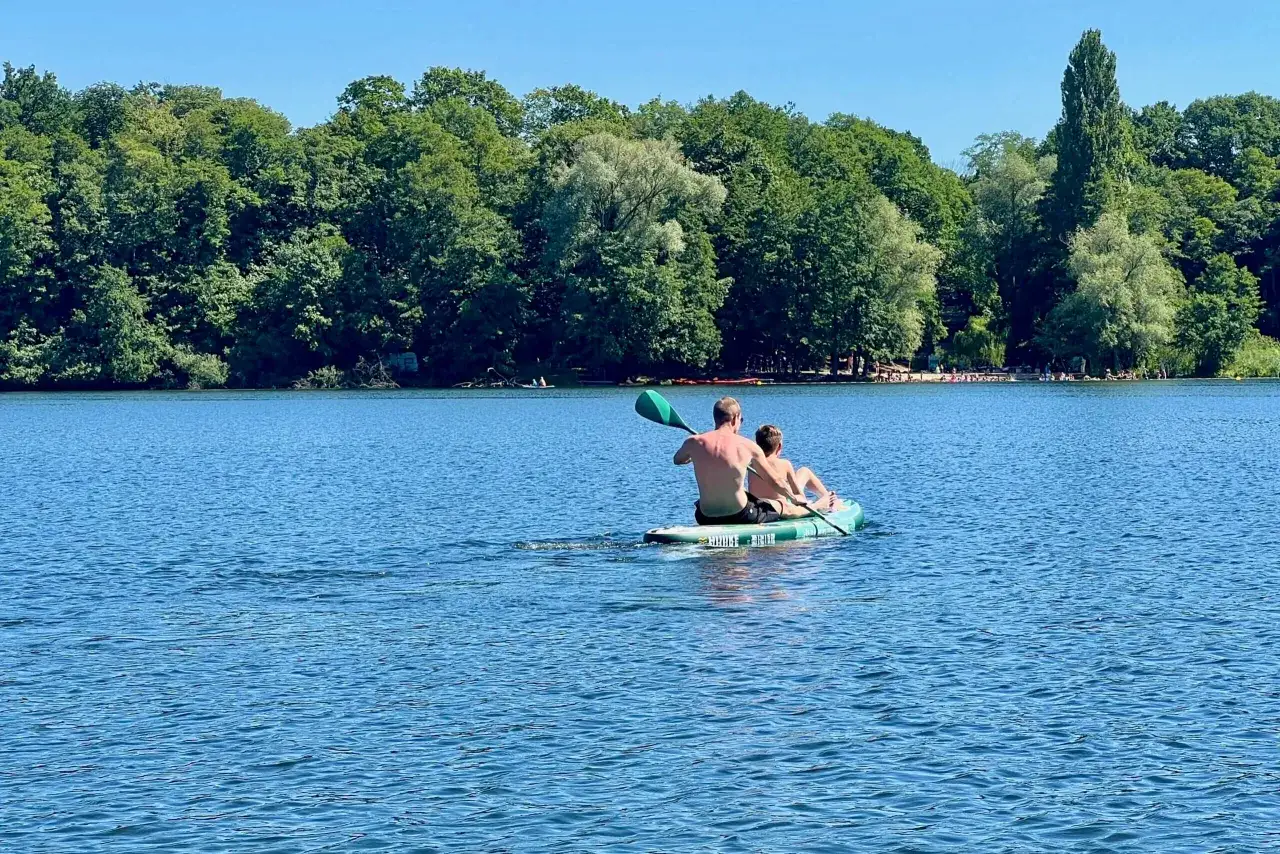 Zwei Personen paddeln auf einem Stand-Up-Board über den glindower See. Im Hintergrund ein dichter grüner Wald und ein blauer Himmel.