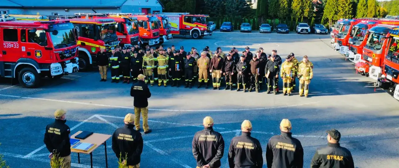 Drużyna ksrg osp pozuje przed rzędem czerwonych wozów strażackich.
