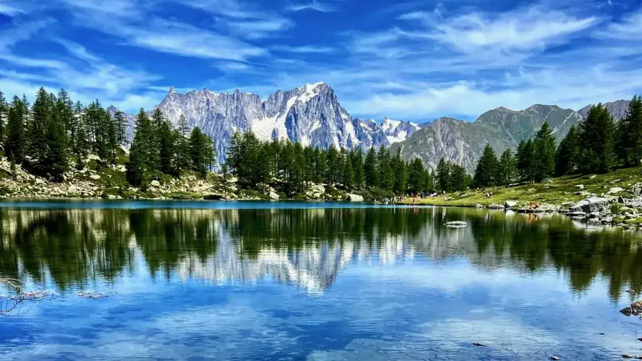Specchio d'acqua cristallino del lago d'Arpy, riflette le maestose montagne innevate e i verdi abeti sotto un cielo azzurro e nuvoloso.