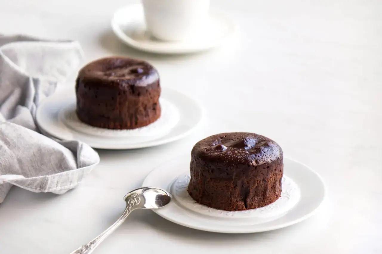Deux fondants au chocolat, moelleux et gourmands, sur des assiettes blanches. Une cuillère argentée et une serviette grise complètent la scène.