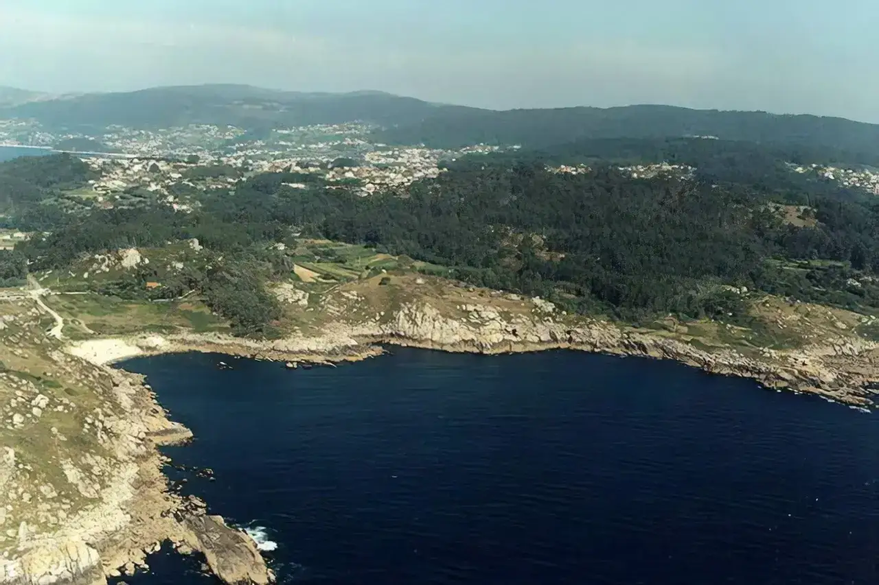 Vista aérea de una costa rocosa con el mar azul profundo. Al fondo, una zona verde con casas y colinas. Se distingue una pequeña playa de ancoradouro.