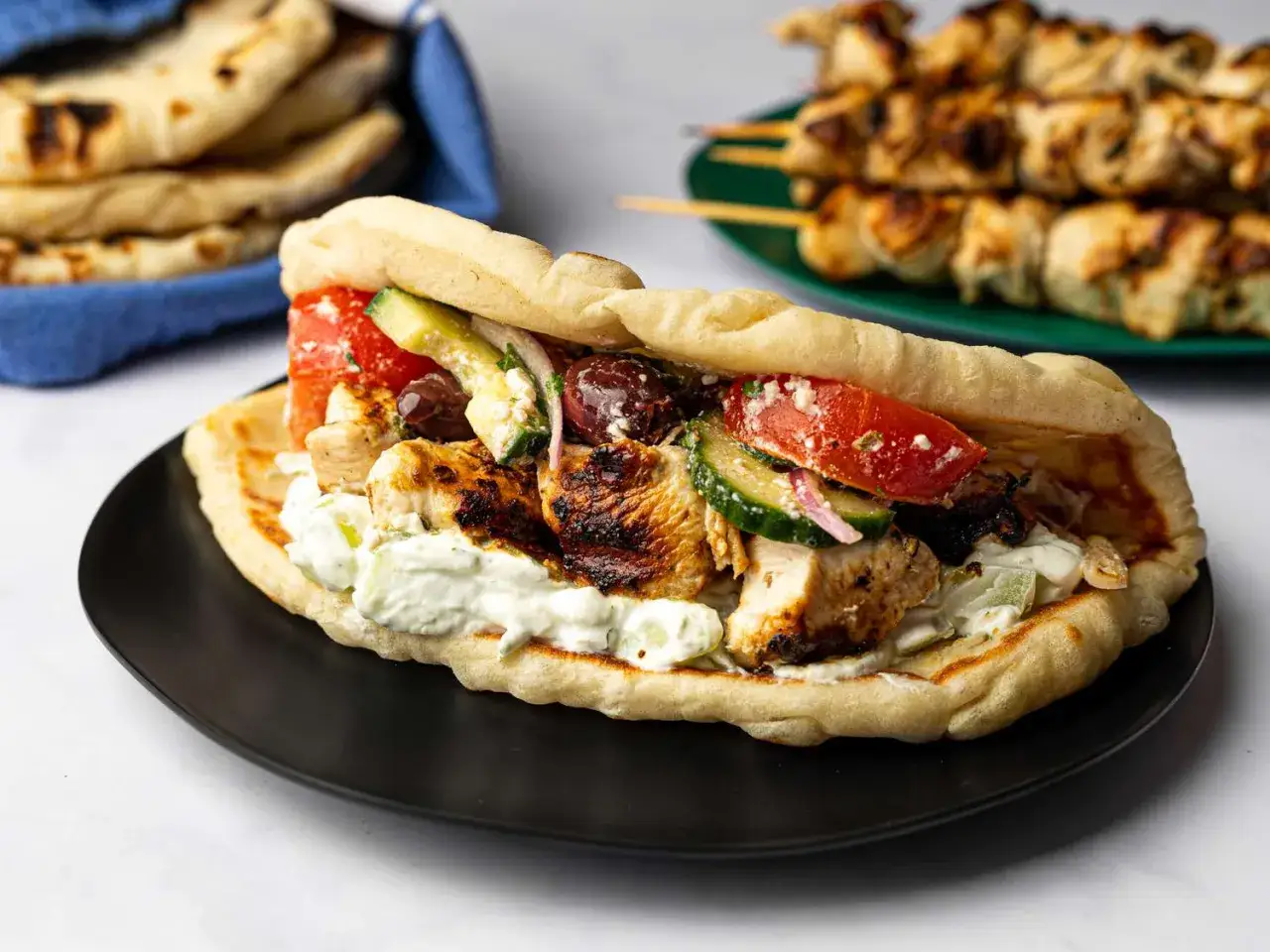 Pyszny gyros przepis: soczysty kurczak, świeże warzywa i sos tzatziki w miękkim picie. Obok pieczone szaszłyki i chlebki pita.