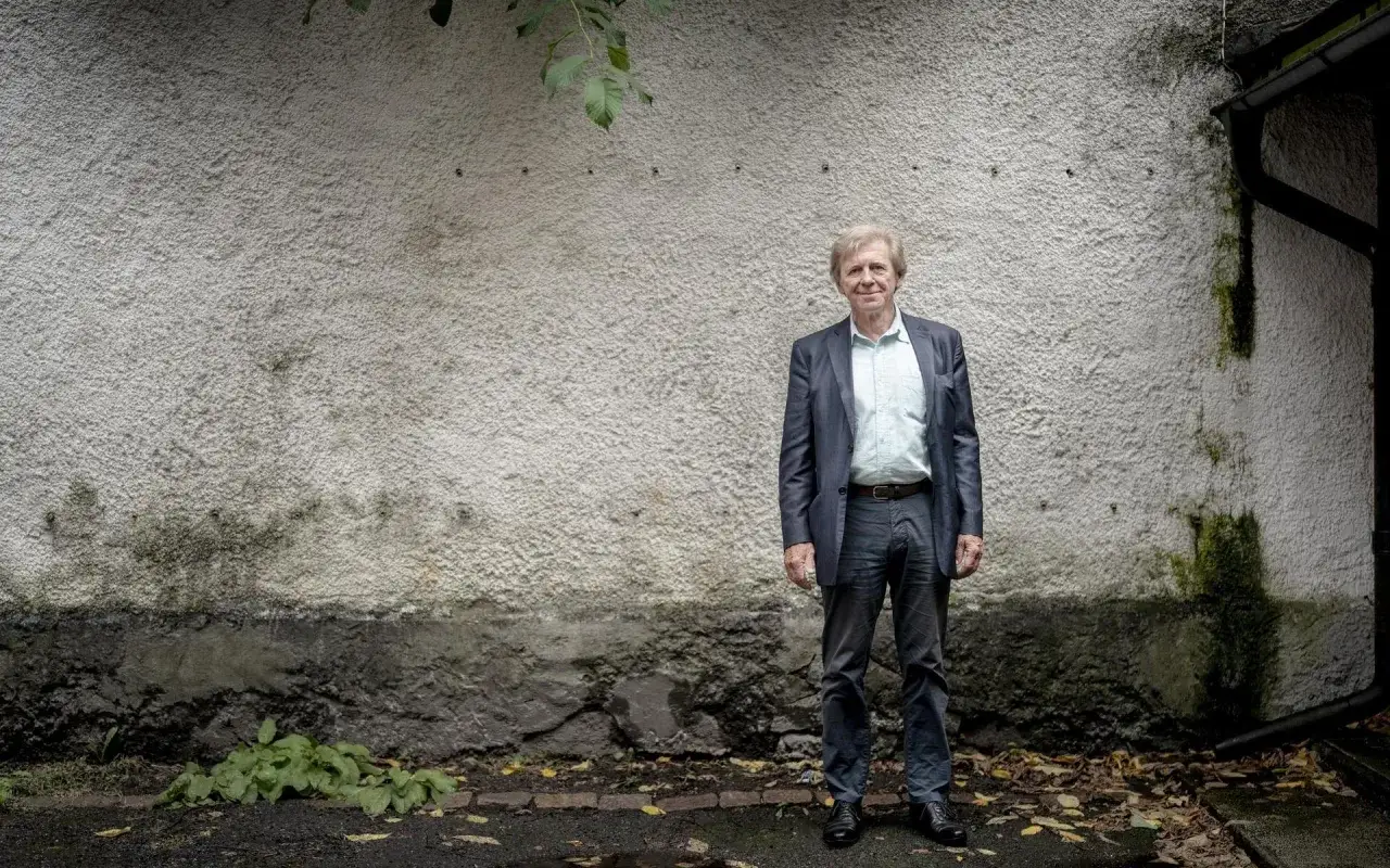 Äldre man i kostym står framför en sliten vägg. Han har bara ben under kostymen.
