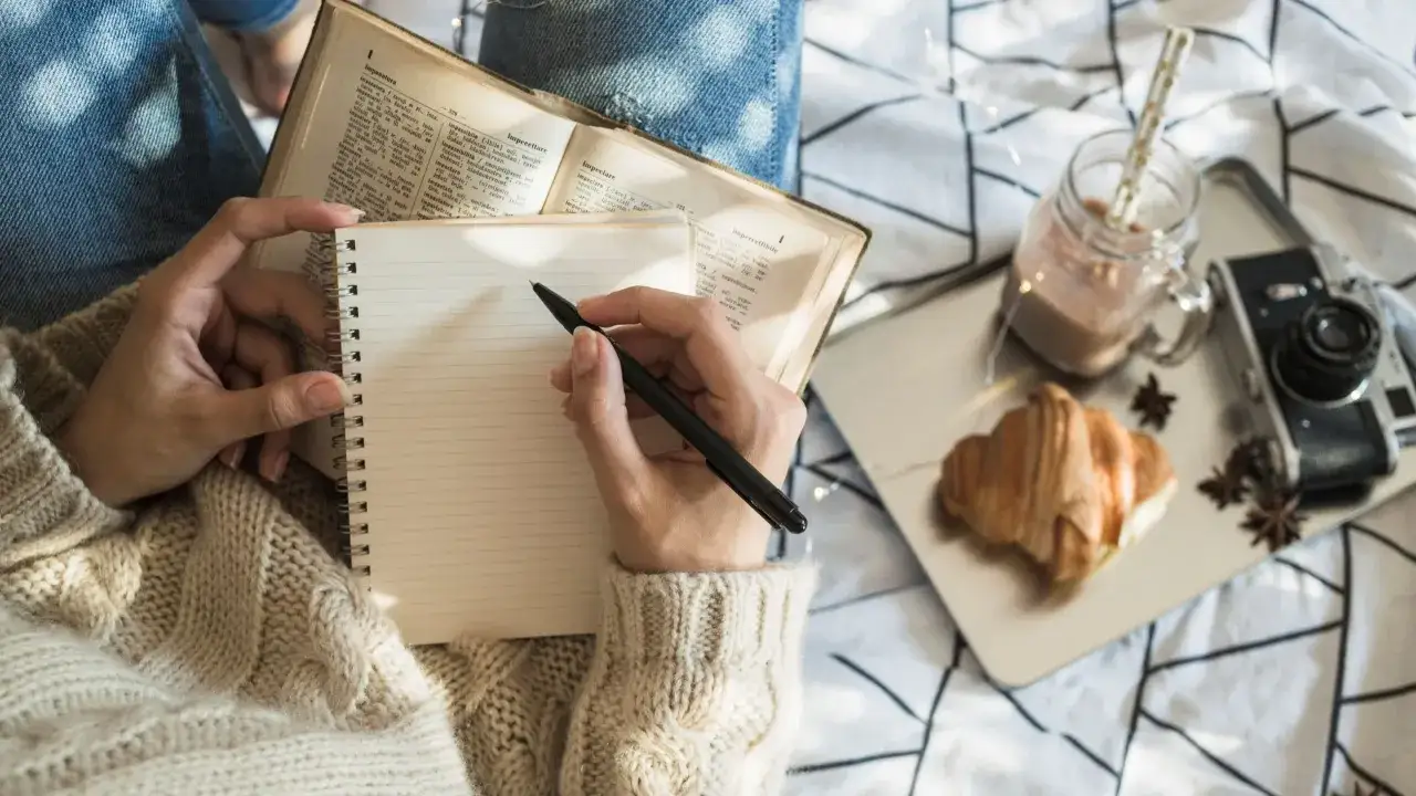 Mani che scrivono su un quaderno, accanto a un libro aperto, croissant, bevanda e macchina fotografica. Un momento per stare bene con se stessi.