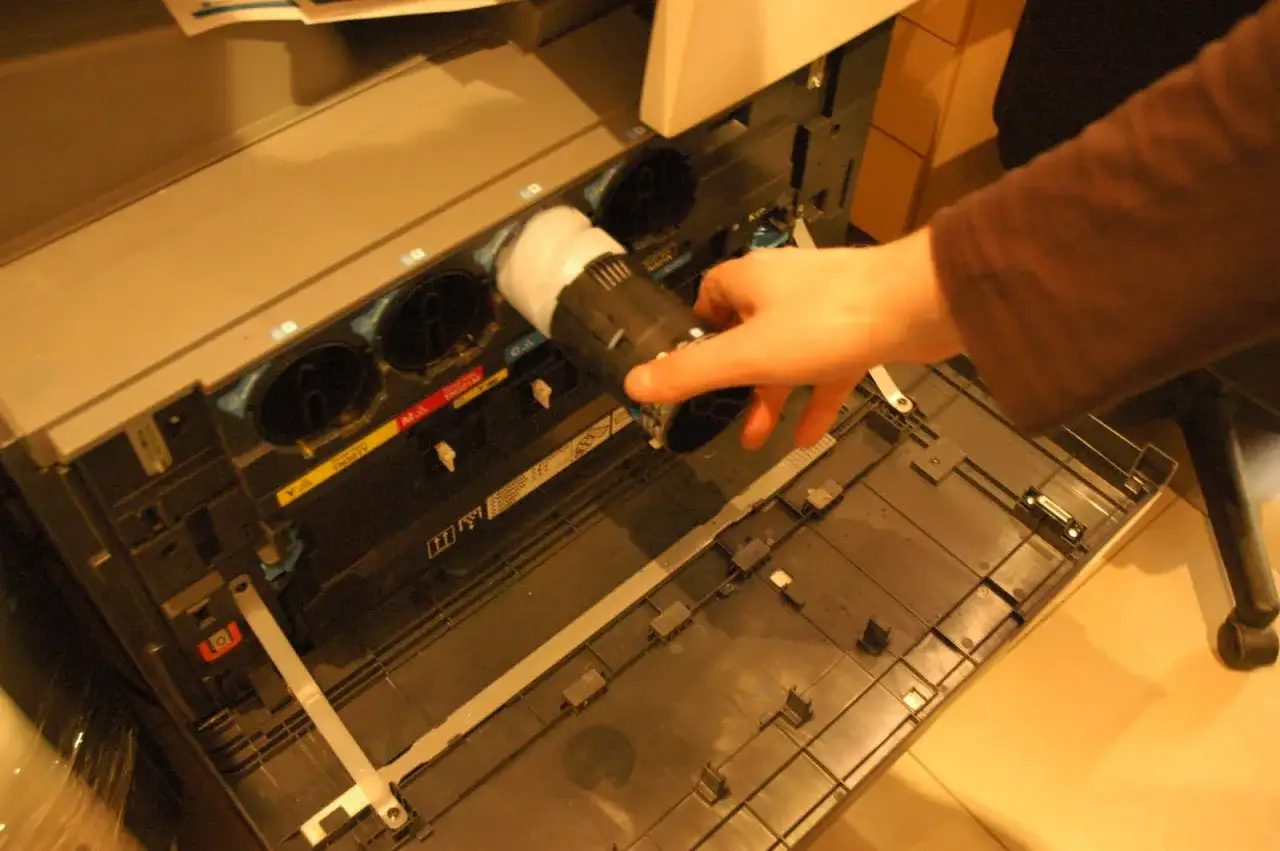 Ręka wymienia toner w drukarce. Widać wnętrze urządzenia z kolorowymi wkładami. Model c203.