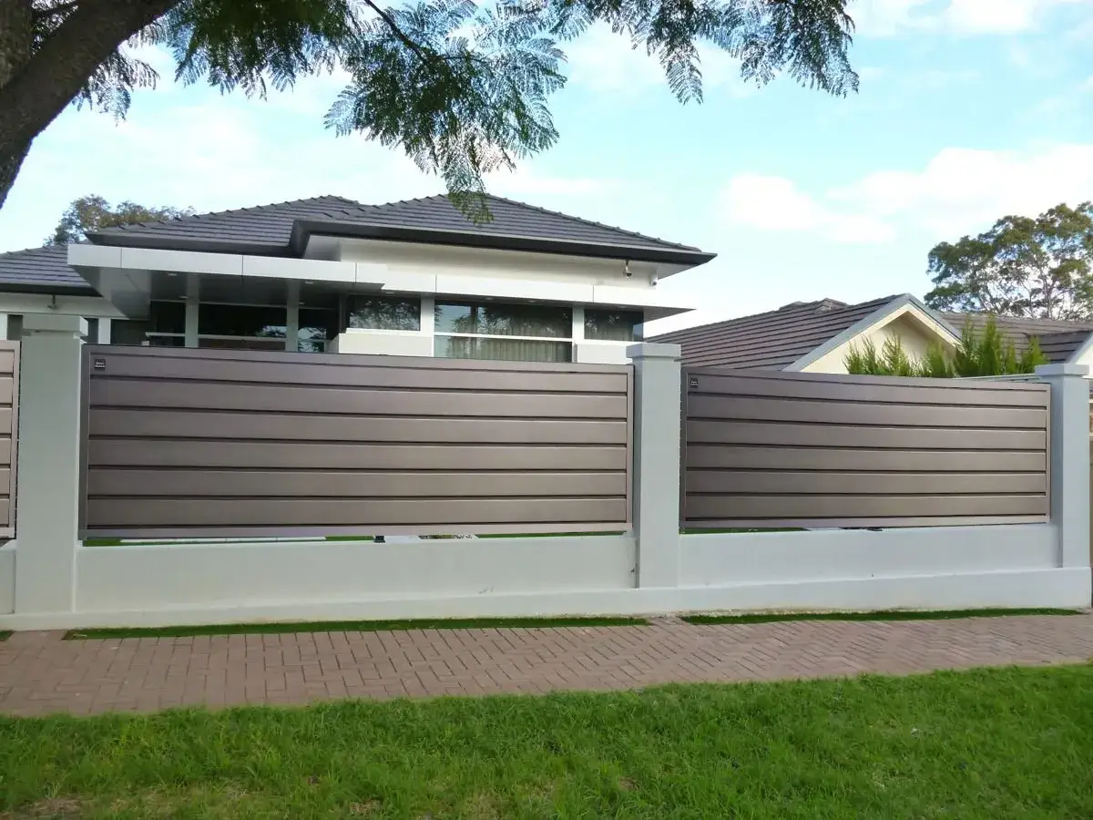 Moderna verja jardin de listones horizontales grises, con pilares blancos y base de hormigón. Casas modernas al fondo.