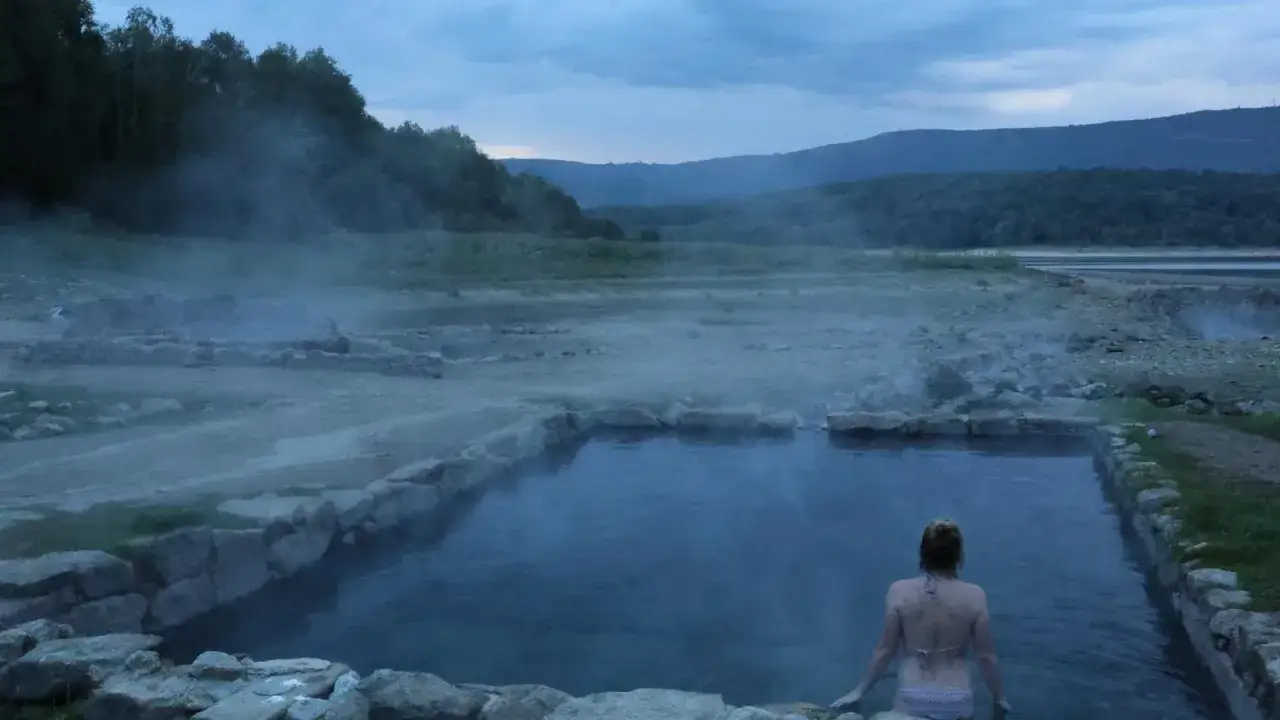 Una mujer se relaja en un estanque de **río caldo** natural, rodeado de rocas y vapor ascendiendo en un paisaje montañoso al anochecer.