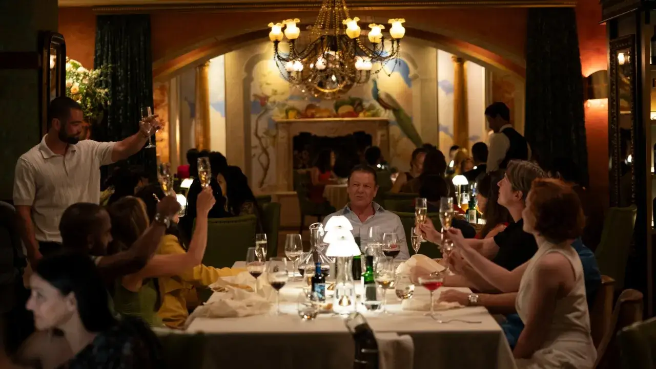 Festliche Stimmung im Restaurant. Gäste stoßen mit Champagner an. Ein Mann hebt sein Glas und sagt: