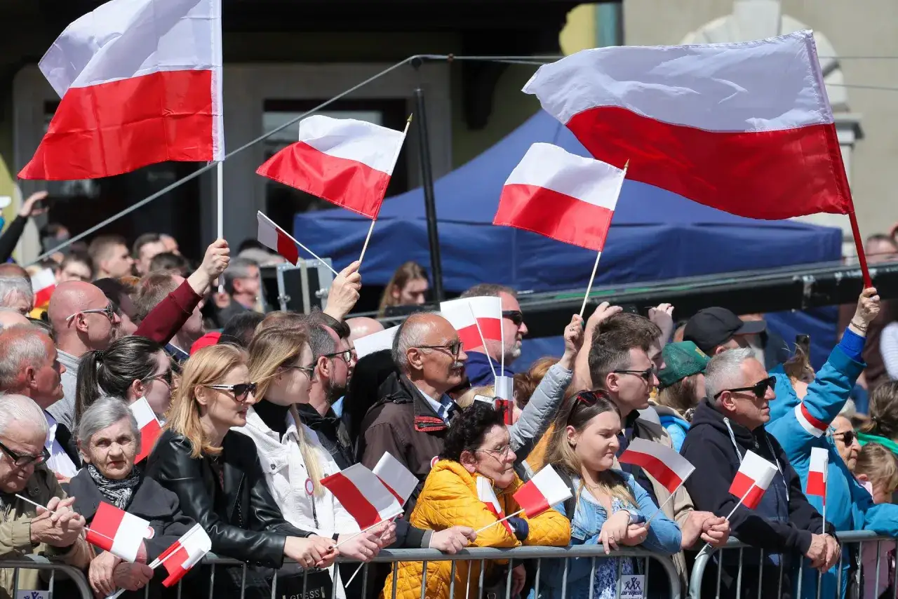 Tłum świętuje obchody 3 maja, machając polskimi flagami. Widać dumę i radość na twarzach ludzi.