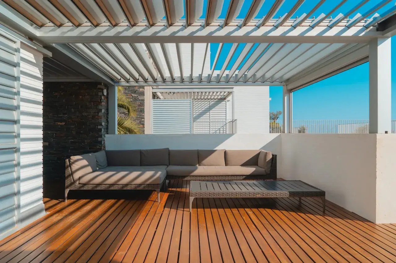 Ideas para decorar un &aacute;tico: un sof&aacute; modular y mesa de centro sobre tarima de madera, bajo una p&eacute;rgola con cielo azul.