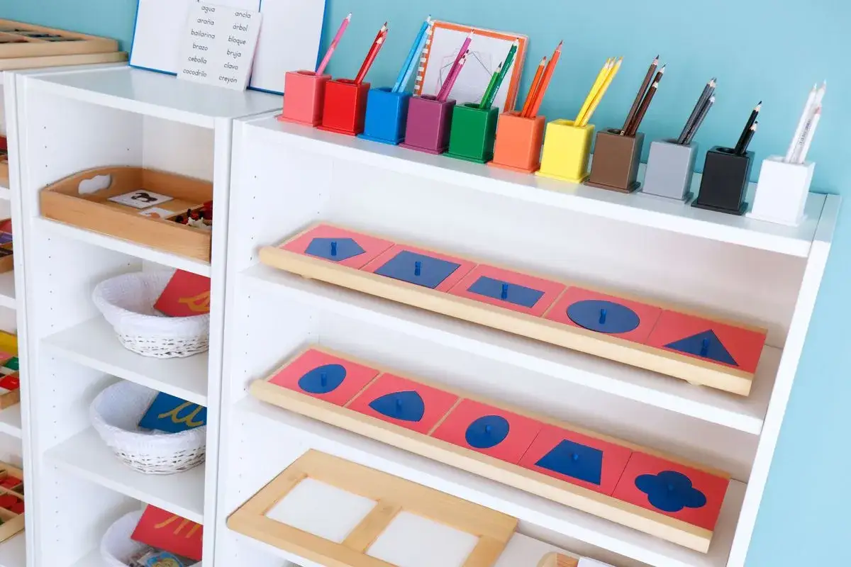 Estanterías con materiales educativos que muestran **qué es Montessori**: encajables de formas geométricas, cestas con tarjetas y portalápices de colores.