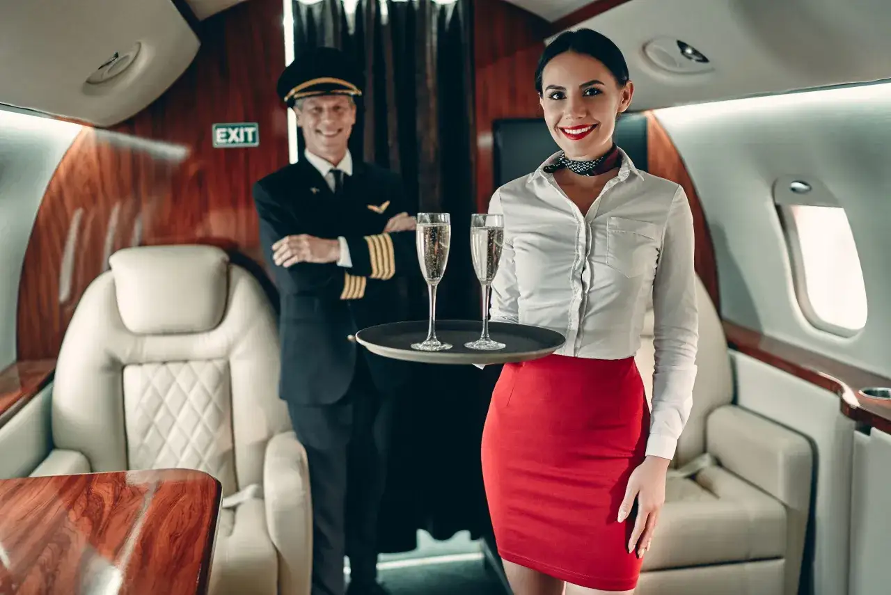 Uśmiechnięta stewardessa w czerwonej spódnicy i białej koszuli niesie tacę z dwoma kieliszkami szampana. W tle pilot w mundurze.
