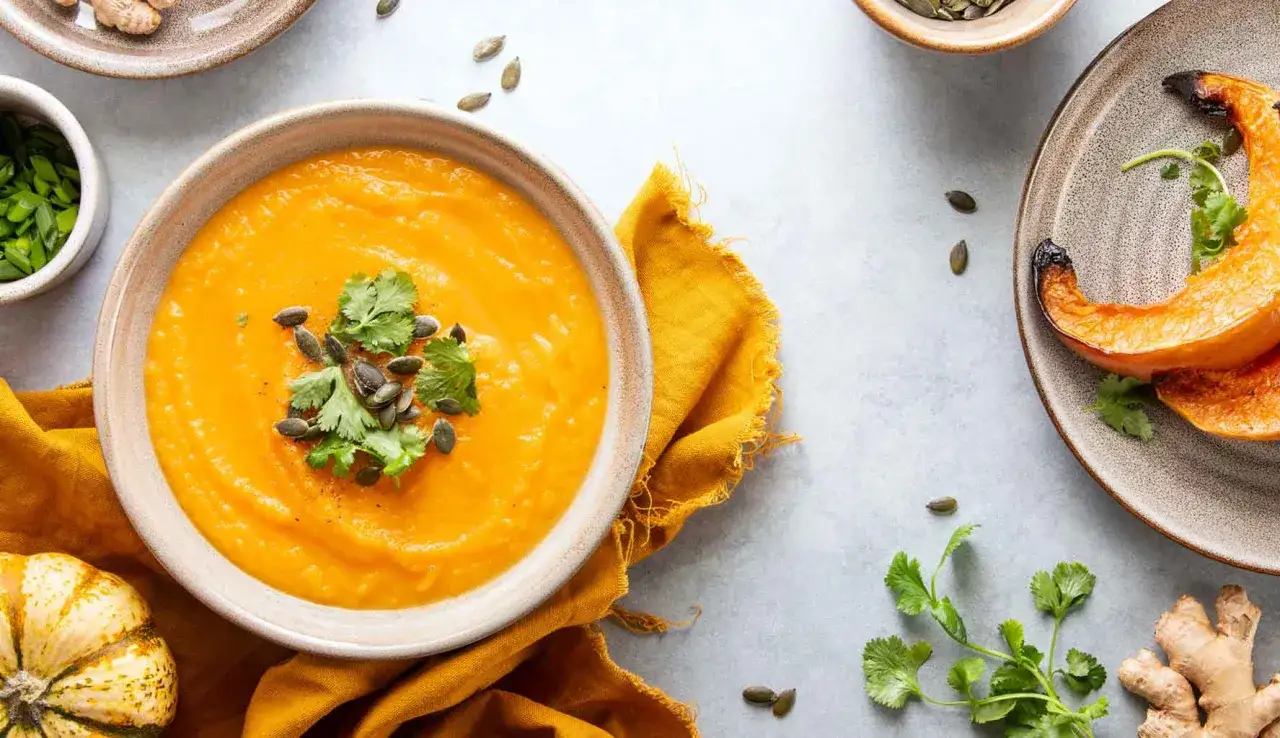 Una ciotola di vellutata di zucca cremosa, guarnita con semi di zucca e coriandolo fresco. Accanto, spicchi di zucca arrostiti e zenzero.
