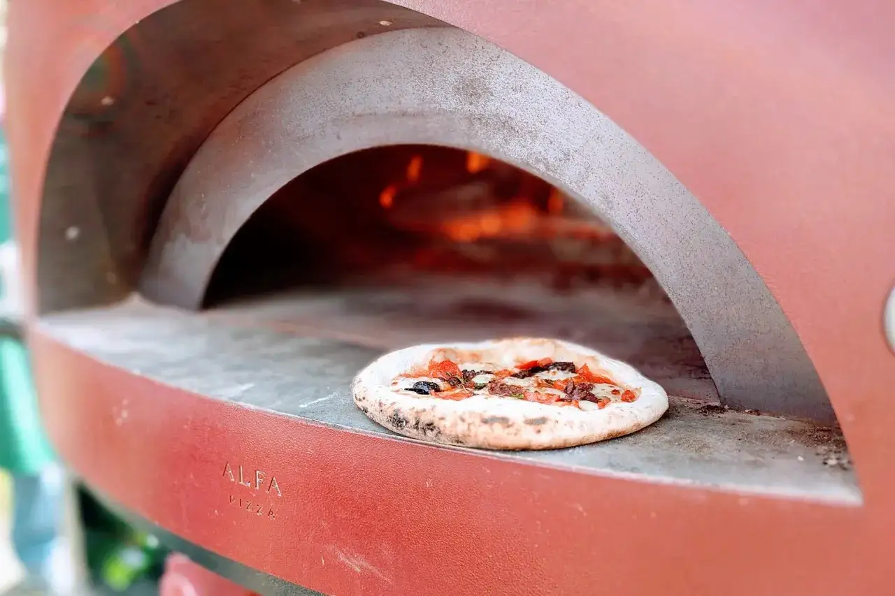 Gotowy do pieczenia placek na tle żarzącego się ognia w piecu. Zastanawiasz się, ile kosztuje piec do pizzy?