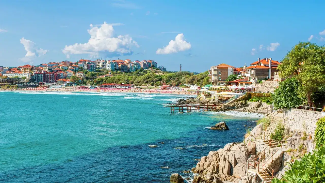 Słoneczna riwiera bułgarska z turkusowym morzem, piaszczystą plażą pełną leżaków i kolorowymi budynkami na wzgórzu.