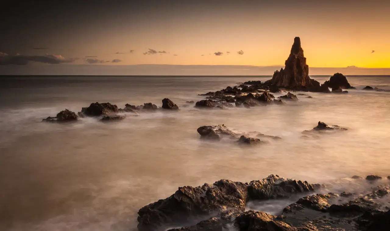 Atardecer en el mirador Cabo de Gata. Rocas escarpadas emergen del mar con aguas sedosas bajo un cielo anaranjado.