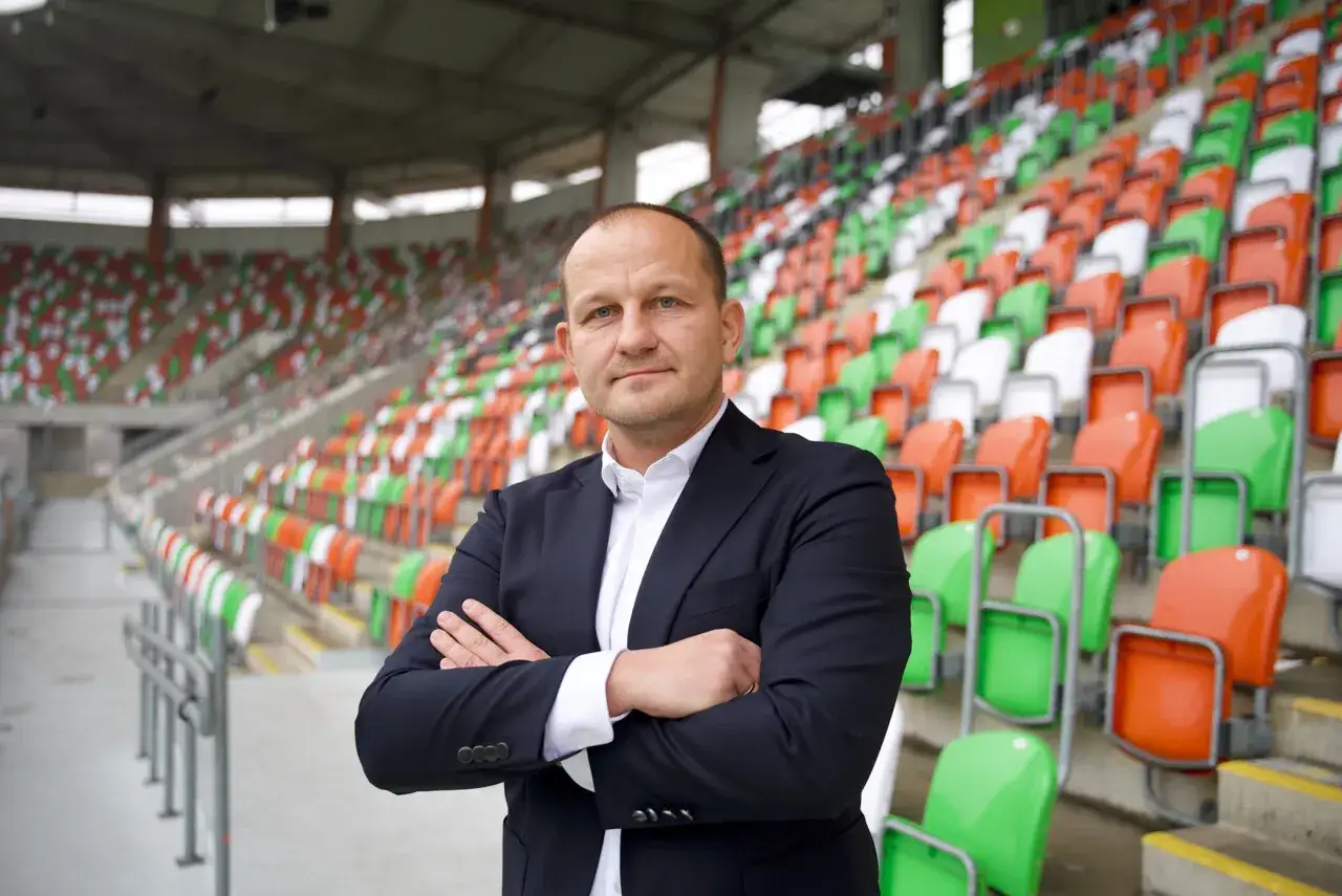 Dyrektor sportowy w garniturze stoi na tle pustych, kolorowych trybun stadionu.