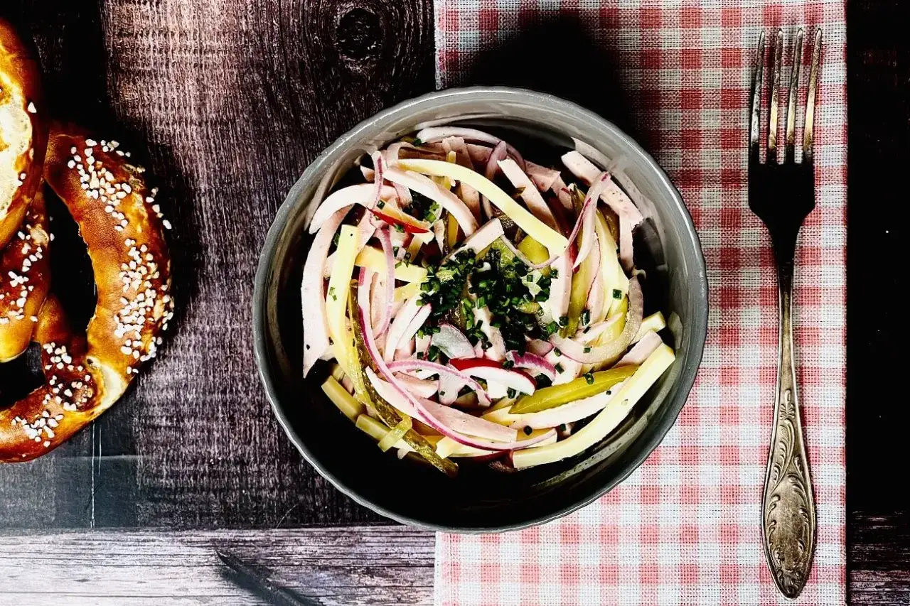 Schmackhafter schweizer wurstsalat mit Zwiebeln und Gurken, garniert mit Schnittlauch. Daneben ein Brezel und eine Gabel.