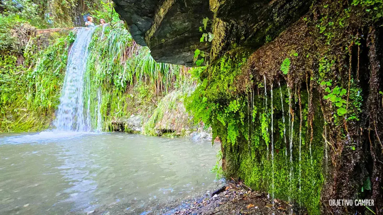 Cascada en la riera del Palau, con rocas cubiertas de musgo y vegetación exuberante. Dos personas disfrutan del agua.