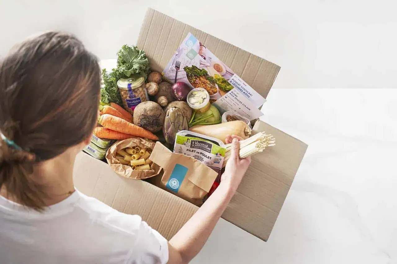 Une personne d&eacute;balle une bo&icirc;te remplie d'ingr&eacute;dients frais : carottes, betteraves, courge, poulet, p&acirc;tes et pois chiches. Id&eacute;al pour un budget courses pour 2 personnes par semaine.