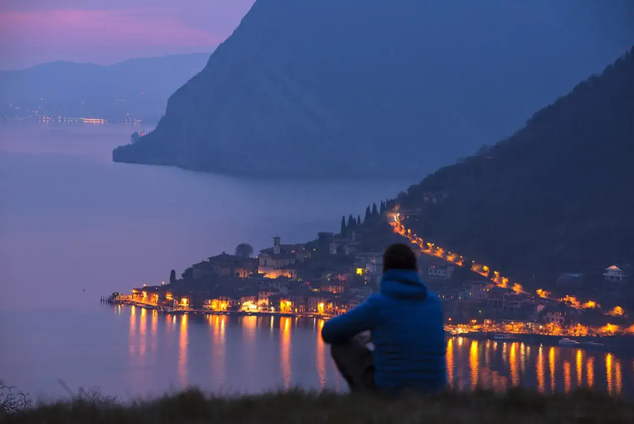 Persona seduta ammira i borghi di Brescia illuminati di notte, con luci che si riflettono sul lago.