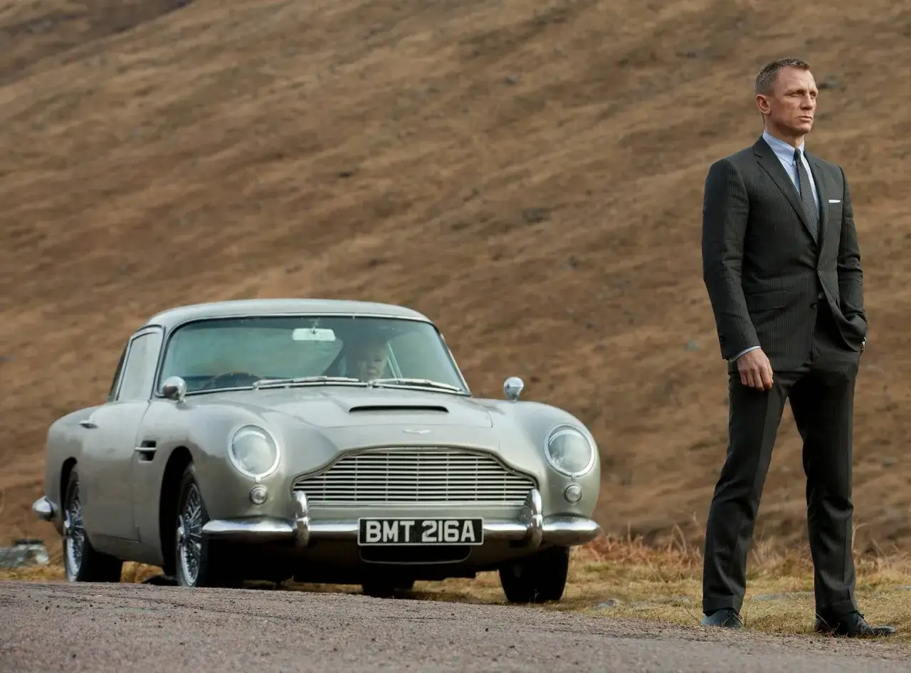 Daniel Craig som James Bond st&aring;r bredvid en Aston Martin DB5, en ikonisk bil fr&aring;n james bond filmer, i en dramatisk skotsk milj&ouml;.